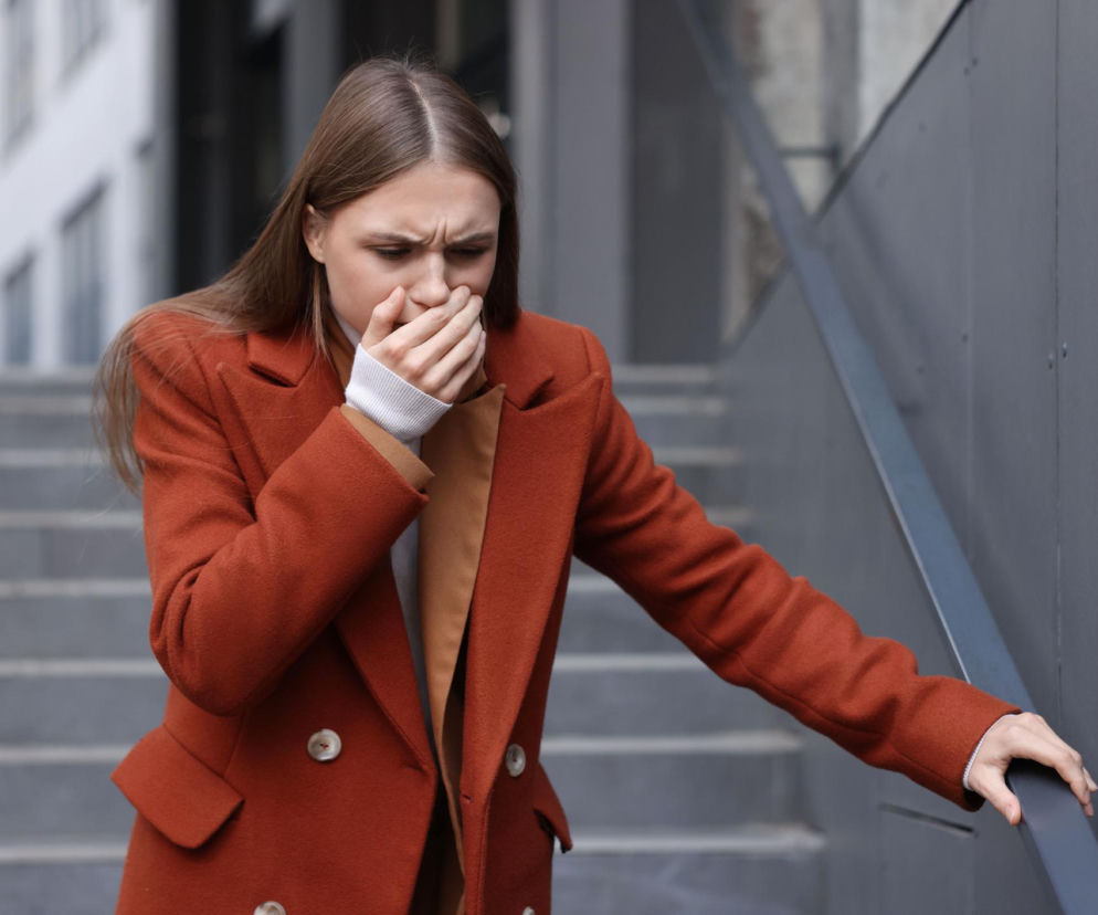 Młoda kobieta w rudym płaszczu z dłonią zasłaniającą usta, opierająca się o poręcz, odczuwająca nudności lub mdłości. Obraz ilustruje objawy norowirusa i zakażenia przewodu pokarmowego, o których można przeczytać na Poradnik Zdrowie.