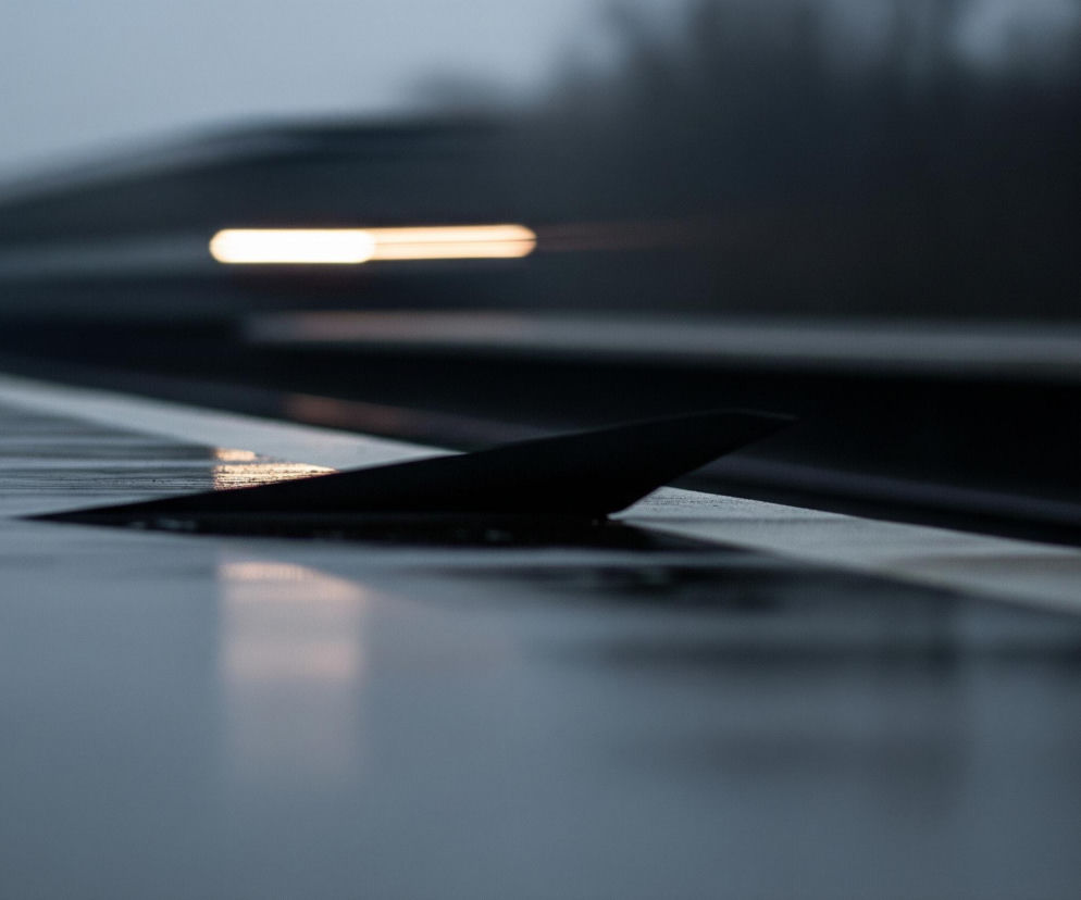 Czarny element o kształcie płetwy wystaje z szarej, błyszczącej powierzchni pokrytej kroplami deszczu, zajmującej dolną połowę obrazu. Powierzchnia ta odzwierciedla rozmazane światła, w tym wyraźną, poziomą, jaskrawożółtą smugę światła w górnej części tła, która zdaje się należeć do szybko poruszającego się obiektu. Tło, rozmyte i niewyraźne, składa się z ciemnych pasów i rozmytych kształtów, sugerujących drogę lub tor. Cała scena utrzymana jest w chłodnych odcieniach szarości i czerni, z jednym, jasnym akcentem koloru.