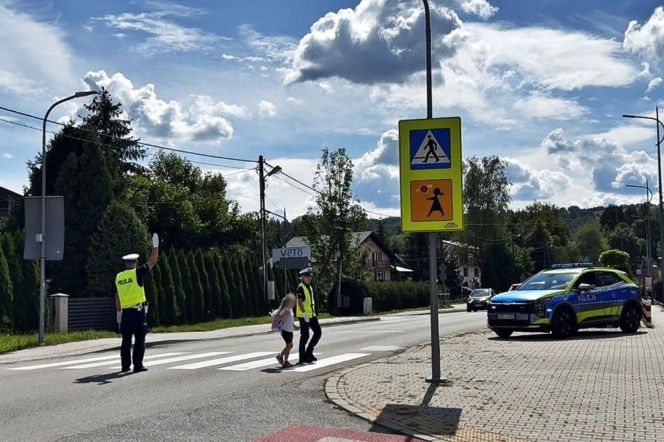 Policjanci pomagają dziecku przejść przez jezdnię przy radiowozie. O akcji policji przeczytasz na Eska Nowy-Sącz.