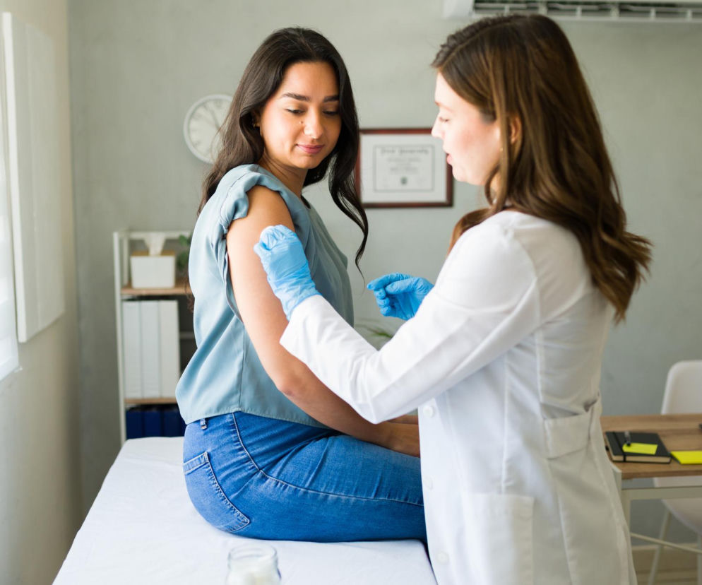 Kobieta w błękitnych jeansach i bluzce siedzi na leżance, otrzymując szczepienie w ramię od lekarki w białym fartuchu i niebieskich rękawiczkach. W tle widoczne okno z żaluzjami i regał. Więcej o bezpieczeństwie szczepionek mRNA przeczytasz na Poradnik Zdrowie.