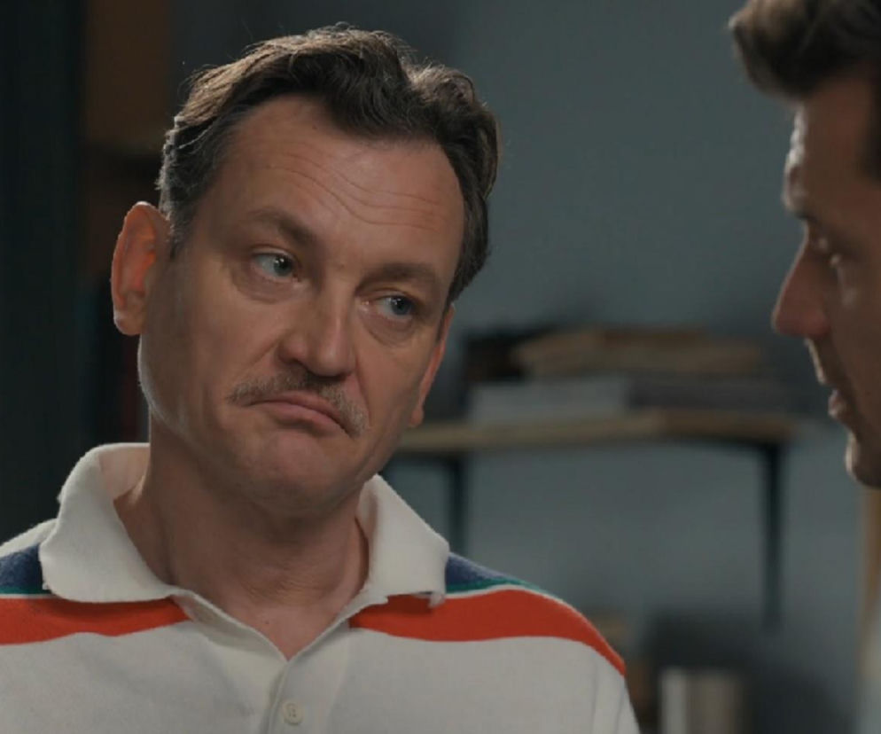 Aktor Paweł Ławrynowicz jako Stefaniak w białej koszulce z kolorowymi paskami, z wąsami i wyraźnym grymasem na twarzy, rozmawia z Darkiem, granym przez Andrzeja Niemyta (widoczny fragment twarzy). Stefaniak próbuje się wykręcić od odpowiedzialności za zniszczenie lusterka, co opisują Super Seriale.