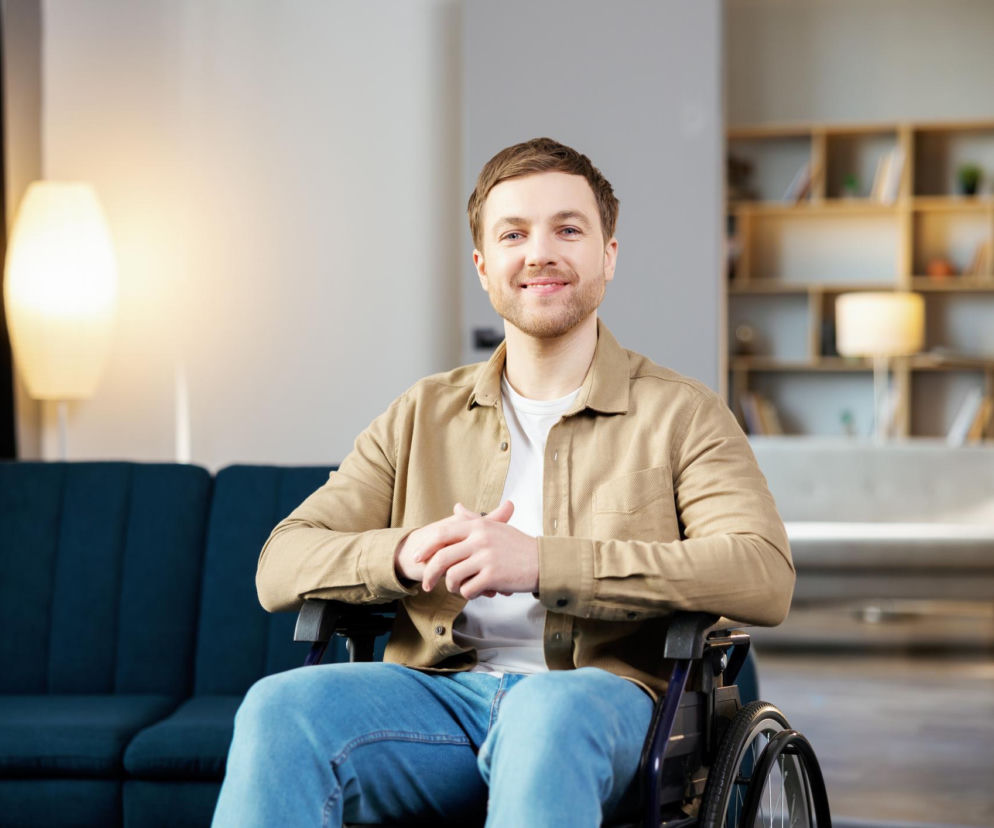 Mężczyzna z zarostem, w beżowej koszuli i jasnych dżinsach, siedzi na wózku inwalidzkim w jasnym salonie, uśmiechając się do kamery. Na portalu Poradnik Zdrowie znajdziesz informacje o ponownym orzeczeniu o niepełnosprawności.