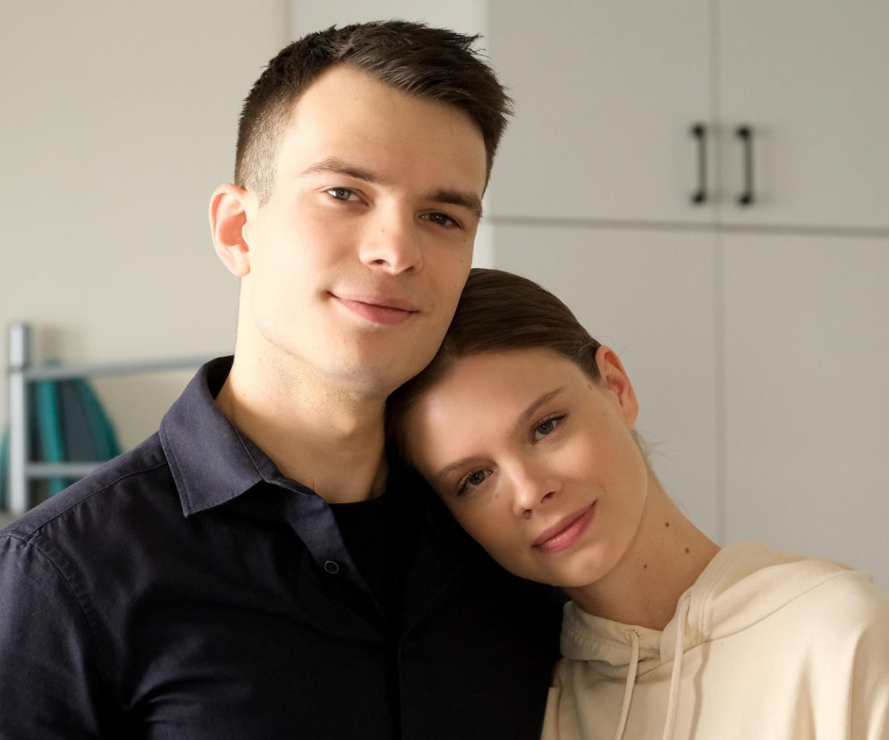 Młoda para, mężczyzna o ciemnych włosach i kobieta z jasną bluzą, obejmująca go w geście czułości. Ich twarze wyrażają spokój i delikatny uśmiech. Zdjęcie ilustruje relacje międzyludzkie, podobne do tych, które można śledzić na portalu Super Seriale.