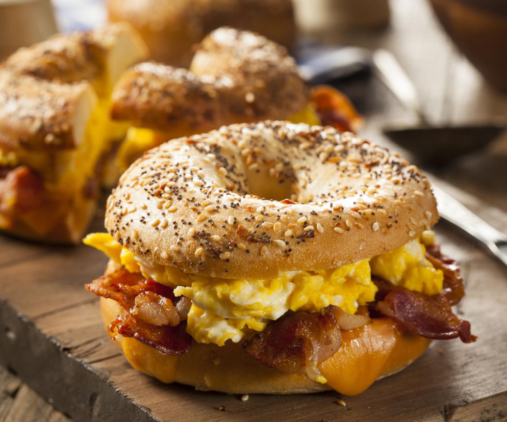 Bajgiel z bekonem, jajecznicą i roztopionym serem cheddar, obficie posypany sezamem i makiem, leży na drewnianej desce. W tle widać kolejnego bajgla. To śniadanie może negatywnie wpływać na serce, o czym przeczytasz na Poradnik Zdrowie.