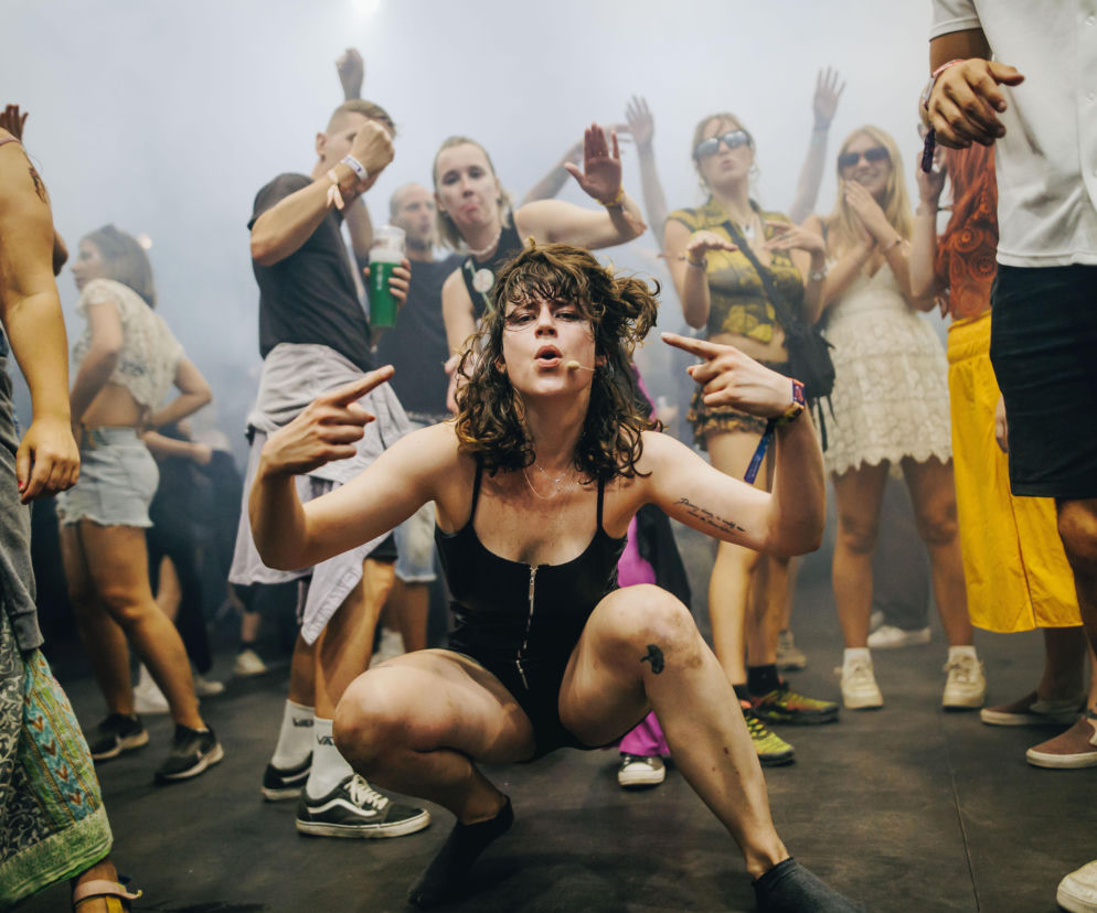 Kobieta w czarnym body kuca i gestykuluje na tle tańczącego tłumu na Sziget Festival. O imprezie przeczytasz w serwisie Eska.