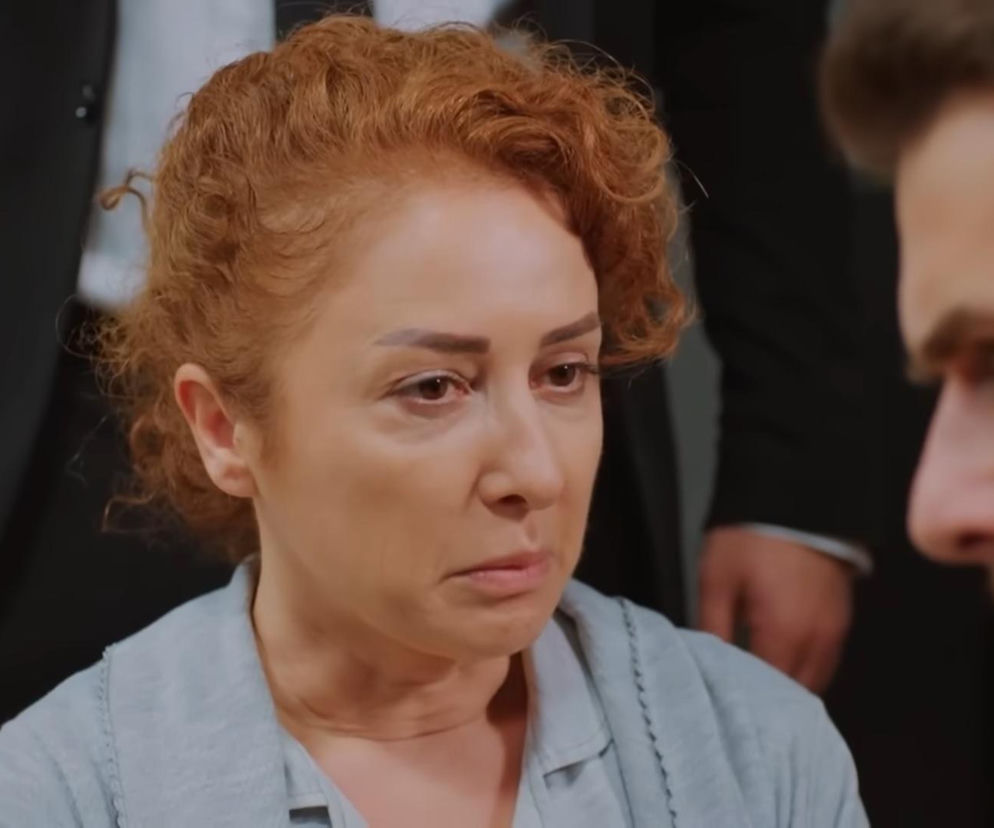 Kobieta z rudymi, kręconymi włosami w szarej bluzce z kołnierzykiem, ze smutnym wyrazem twarzy spogląda na niewyraźną postać mężczyzny, widoczną w prawym rogu. W tle majaczy fragment ciemnego garnituru, co sugeruje formalne spotkanie. Więcej o losach bohaterów na Super Seriale.