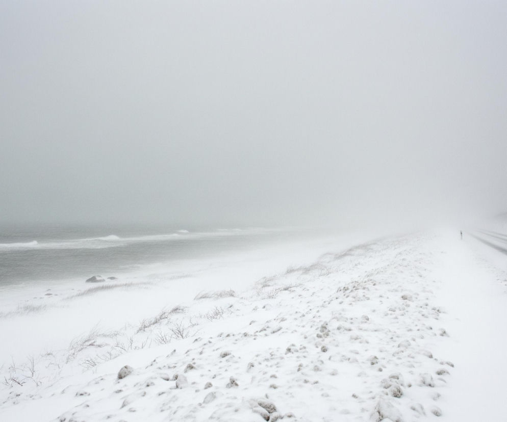 Na pierwszym planie rozciąga się ośnieżone pobocze, pokryte nierównymi grudami śniegu i wystającymi, nagimi gałązkami krzewów. Po lewej stronie widać wzburzone morze z białymi falami, a linia brzegowa zlewa się z horyzontem w odcieniach szarości. Po prawej stronie biegnie droga z ciemnym asfaltem, częściowo przysypana śniegiem, która znika w dali w mlecznej mgle, a obok niej rośnie pas ciemnych drzew lub krzewów. Całość otacza jasnoszare, zamglone niebo, sprawiając wrażenie zimowej, monotonnej pogody.