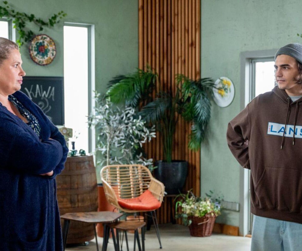 Aldona, w ciemnoniebieskim kardiganie, stoi ze skrzyżowanymi rękami, patrząc na Romea w brązowej bluzie z kapturem i szarej czapce. Romeo stoi z rękami na biodrach, odwrócony nieco od Aldony. Scena przedstawia ich w kawiarni. Więcej o ich relacji w serialu „Barwy szczęścia” znajdziesz na portalu Super Seriale.