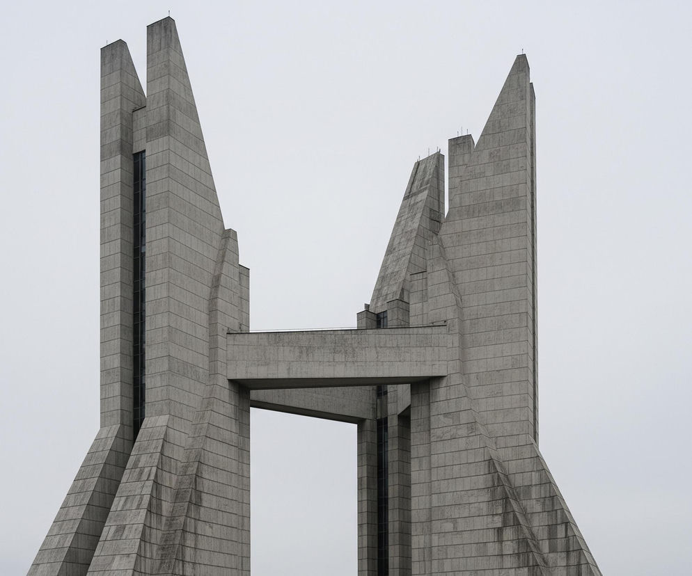 Centralny element obrazu to monumentalna, betonowa konstrukcja architektoniczna o symetrycznym kształcie, składająca się z dwóch strzelistych wież połączonych na wysokości poziomu w górnej części. Wieże mają skośne, geometryczne płaszczyzny i są zbudowane z kwadratowych lub prostokątnych płyt betonu, z widocznymi poziomymi i pionowymi fugami. Na lewej wieży znajduje się pionowy pas ciemnych okien lub szczelin, biegnący wzdłuż jej wewnętrznej krawędzi. Górna część wież jest zakończona ostrymi, spiczastymi wierzchołkami, a na szczytach widać niewielkie, pionowe elementy przypominające anteny. Cała konstrukcja utrzymana jest w szaro-popielatej kolorystyce, z subtelnymi odcieniami brudu i zacieków na powierzchni betonu. Tło stanowi jednolite, jasnoszare niebo, co dodatkowo podkreśla surowość i monumentalność budowli.