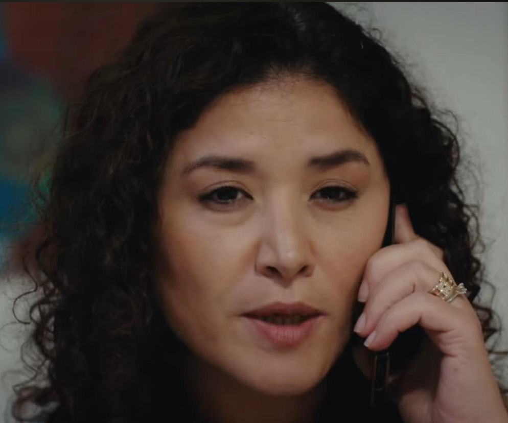 Kobieta z ciemnymi, kręconymi włosami i pierścionkiem na palcu, trzymająca telefon przy uchu, rozmawia z poważnym wyrazem twarzy. Jej mina sugeruje konsternację lub zaniepokojenie, co doskonale oddaje emocje związane z intrygami Cansel w serialu Dziedzictwo, o których przeczytasz na Super Seriale.