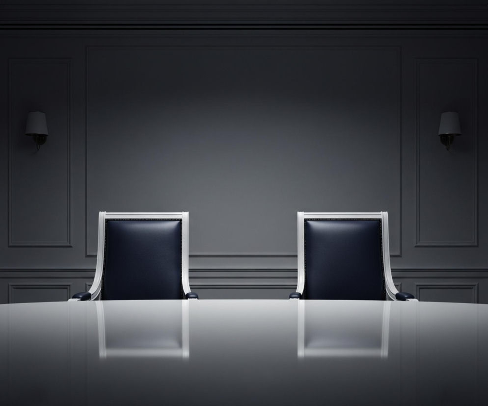 Dwa identyczne, puste krzesła z ciemnogranatową tapicerką i białymi ramami stoją przy dużym, owalnym stole z błyszczącym, jasnym blatem, odbijającym ich kształt. Pomieszczenie charakteryzuje się ciemnymi, panelowymi ścianami z subtelnymi ozdobnymi listwami, a po obu stronach centralnej części ściany znajdują się drzwi oraz kinkiet. Całość utrzymana jest w stonowanych odcieniach szarości i błękitu, z miękkim oświetleniem tworzącym wrażenie intymności.