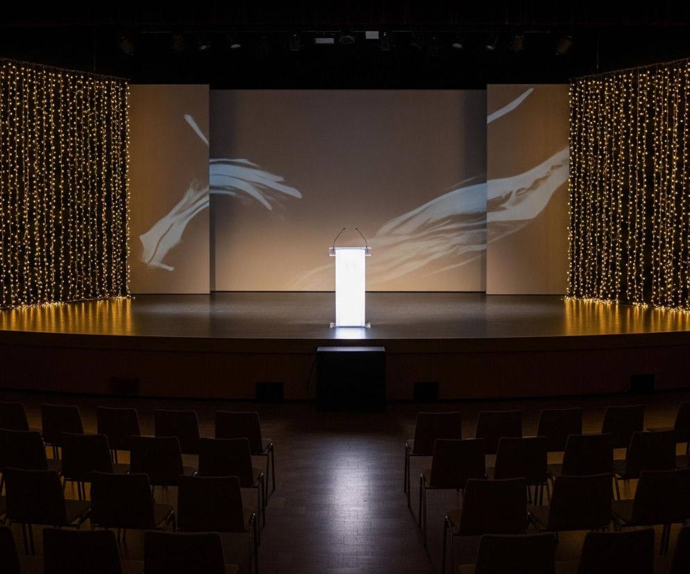 Na pierwszym planie widoczne są rzędy pustych krzeseł dla publiczności w ciemnej sali. Jasno oświetlona scena znajduje się na dalszym planie, gdzie centralnie stoi biała, podświetlana mównica z mikrofonami. Za mównicą rozciąga się duży ekran, na którym wyświetlane są abstrakcyjne kształty w odcieniach bieli i jasnego błękitu na beżowo-szarym tle. Po obu stronach ekranu wiszą kurtyny z migoczących złotych światełek, które odbijają się w błyszczącej podłodze sceny.