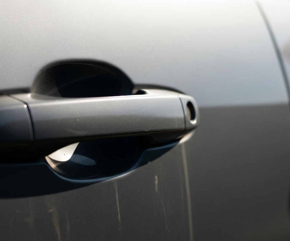 Klamka samochodowa w szarym, metalicznym kolorze zajmuje centralną część obrazu. Klamka ma aerodynamiczny kształt i odblaskową powierzchnię, częściowo oświetloną silnym światłem, które tworzy wyraźne cienie pod nią i w zagłębieniu uchwytu. Tło to rozmyta karoseria pojazdu, również w odcieniach szarości, z widocznymi refleksami światła i drobnymi cząsteczkami kurzu.