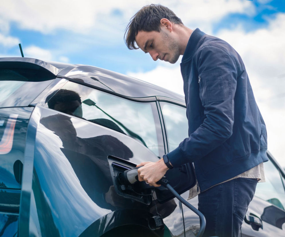Mężczyzna podłączający kabel do gniazda ładowania samochodu elektrycznego. Kierowca ładuje auto na stacji ładowania, korzystając z nowej, niższej stawki. O ekonomicznych zmianach dla posiadaczy elektryków przeczytasz na Super Biznes.