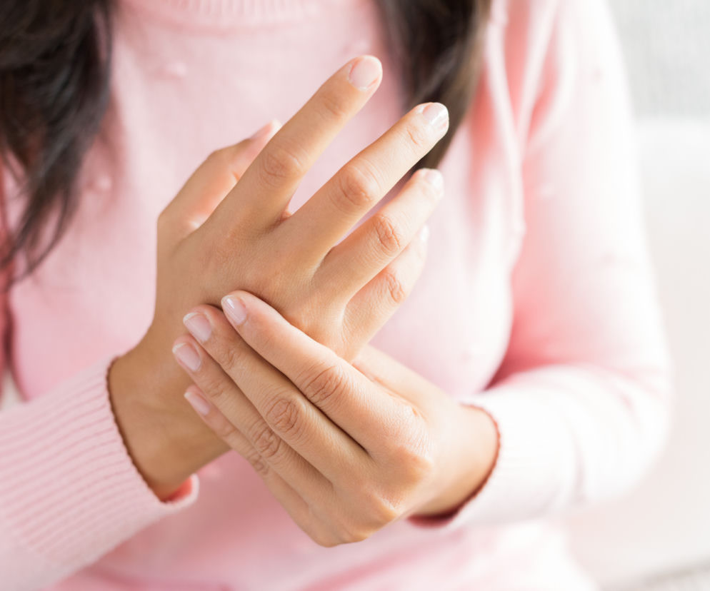 Kobieta masuje nadgarstek dłonią, odczuwając ból lub mrowienie. To typowy objaw przeciążenia rąk po pracy przy komputerze, który może świadczyć o zespole cieśni nadgarstka. O przyczynach i sposobach łagodzenia dolegliwości przeczytasz na Poradniku Zdrowie.