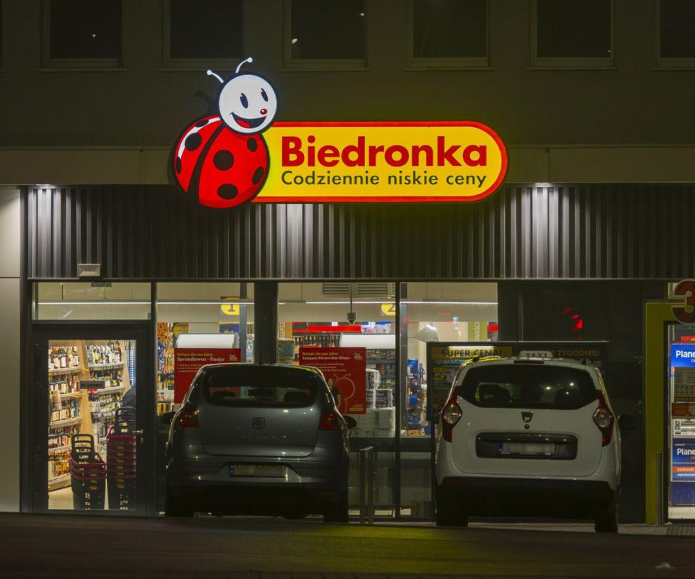 Nocne ujęcie wejścia do sklepu Biedronka z widocznym logotypem i hasłem „Codziennie niskie ceny”. Przed wejściem dwa zaparkowane samochody. Zdjęcie ilustruje wydłużone godziny otwarcia Biedronki przed świętami, o czym czytać można na portalu Super Biznes.