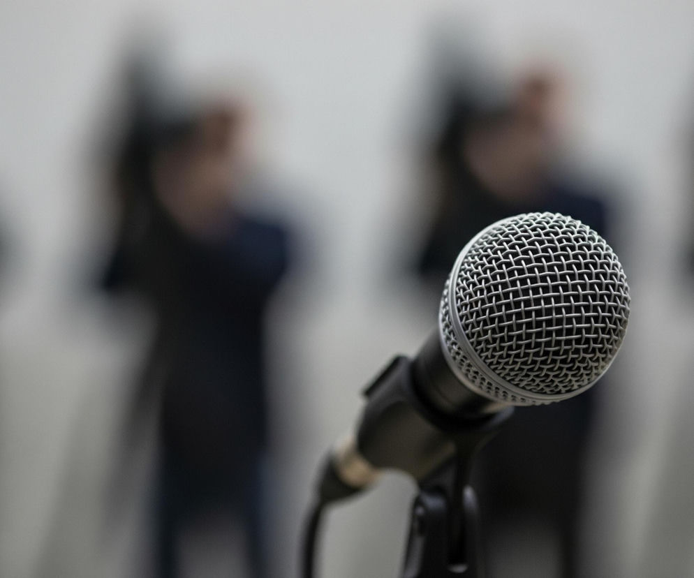 Centralnym elementem obrazu jest mikrofon w kolorze srebrnym i czarnym, umieszczony na statywie, widoczny w dolnej części kadru i lekko skierowany w prawo. Głowica mikrofonu, pokryta metalową, siatkową osłoną, jest ostra i dobrze widoczna, podobnie jak część jego czarnego korpusu i uchwytu statywu. Tło za mikrofonem jest rozmyte, przedstawiając niewyraźne sylwetki kilku osób w ciemnych ubraniach, stojących w rzędzie. Dominują odcienie szarości i czerni, z jaśniejszymi fragmentami tła między postaciami.