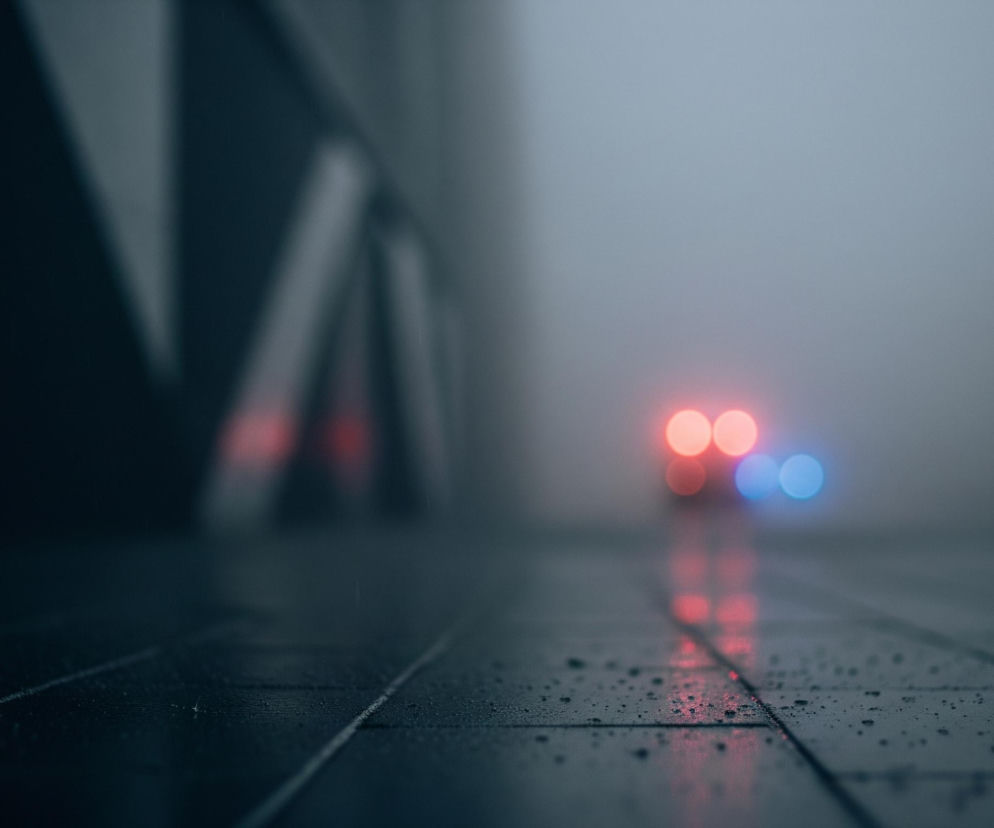 Ulica pokryta jest kroplami deszczu, które odbijają światła. W oddali, w rozmazanym tle, widoczne są cztery rozmyte punkty świetlne: trzy czerwone i jeden niebieski, z czerwoną poświatą odbijającą się na mokrej nawierzchni. Po lewej stronie kompozycji znajduje się ciemny, niewyraźny kształt przypominający filar lub element architektoniczny, częściowo zanurzony w półcieniu. Całość utrzymana jest w chłodnej tonacji szarości i granatu, z wyraźnym efektem głębi ostrości.