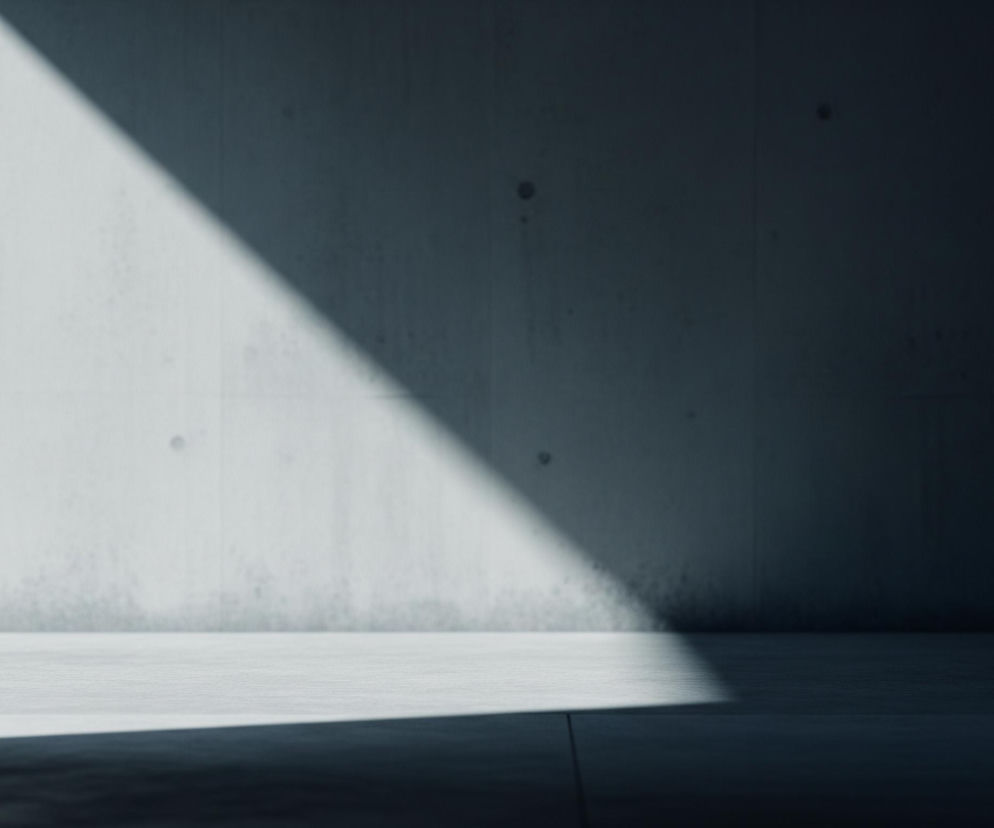 Wnętrze minimalistycznego pomieszczenia z surowych, betonowych ścian i podłogi. Światło słoneczne pada przez niewidoczne okno, tworząc ukośny, jasny pas na lewej ścianie i fragment podłogi. Reszta wnętrza jest pogrążona w półmroku, z wyraźnym przejściem tonalnym od jasnej szarości do ciemnego granatu. Na lewej ścianie widoczne są cztery ciemniejsze okrągłe punkty, prawdopodobnie ślady po szalunku, a także poziome i pionowe linie podziału.