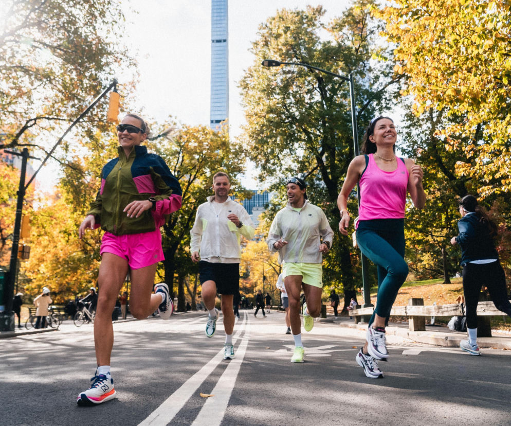 Grupa uśmiechniętych biegaczy w kolorowych strojach sportowych, trenujących na tle jesiennego parku i miejskiego krajobrazu. Promocja wydarzenia New Balance Run House dla miłośników biegania. Więcej o aktywnościach dla biegaczy przeczytasz na Super Biznes.
