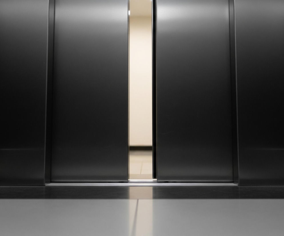 Widoczne są otwierające się drzwi windy w kolorze ciemnoszarym, z wąską pionową szczeliną pośrodku, przez którą przebija jasne, beżowe światło z korytarza. Po obu stronach drzwi rozciągają się ciemne ściany szybu windy. Podłoga w kabinie windy jest błyszcząca, grafitowa, z wyraźnym odbiciem światła i cienia od drzwi.