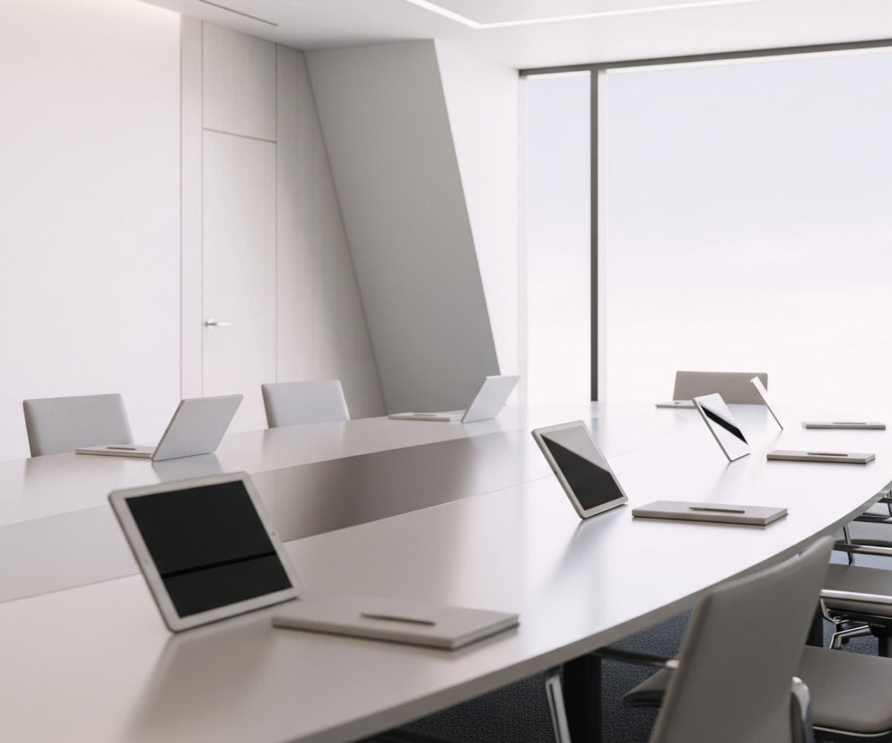 Centralnym elementem jest długi, biały stół konferencyjny w kształcie elipsy, otoczony krzesłami biurowymi na kółkach. Na stole rozmieszczone są laptopy i tablety z czarnymi ekranami, a także płaskie, prostokątne notesy w jasnych odcieniach szarości. Pomieszczenie charakteryzuje się minimalistycznym designem z białymi ścianami, jedną z nich zajmuje wysokie, prostokątne okno z widokiem na jasne, rozmyte niebo. Podłoga pokryta jest ciemnoszarym dywanem.