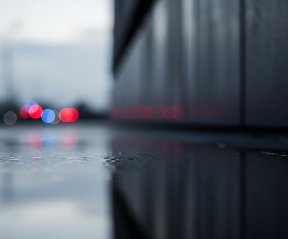 Pierwszy plan ukazuje mokrą, gładką powierzchnię, prawdopodobnie asfaltu lub chodnika, odbijającą światła. W prawym górnym rogu znajduje się fragment ciemnej ściany budynku o teksturowanej powierzchni, z widocznymi poziomymi liniami. W tle, rozmyte czerwone i niebieskie światła tworzą efekt bokeh, sugerując obecność pojazdów lub sygnalizacji świetlnej. Całość utrzymana jest w chłodnych odcieniach szarości i błękitu, z akcentami czerwieni, co nadaje scenie nocny lub zmierzchowy charakter.