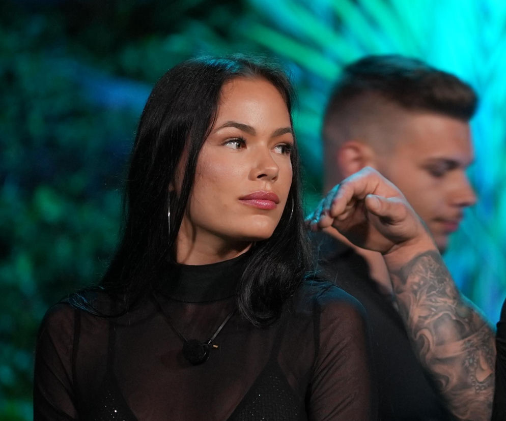 Magda z programu Hotel Paradise w prześwitującej bluzce i czarnej spódnicy, siedząca obok Andrzeja w czarnej koszuli. W tle palmy. Na portalu Super Seriale przeczytasz o trudnym dzieciństwie Magdy.