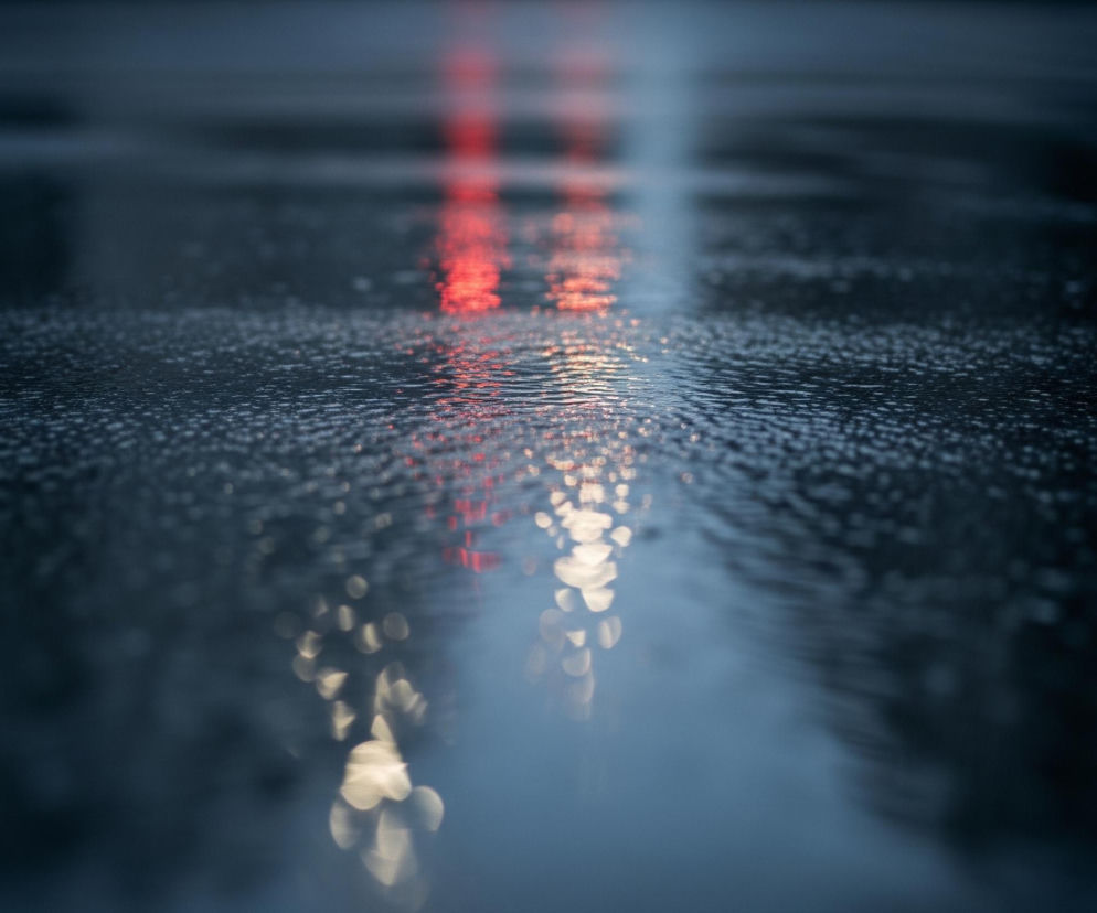 Mokra, ciemna nawierzchnia jezdni rozciąga się od dołu obrazu, zlewając się z rozmytym tłem. Powierzchnia ta, pokryta drobnymi, błyszczącymi kroplami wody, odbija rozmazane światła, tworząc podłużne smugi. Na środku obrazu widoczne są intensywnie czerwone refleksy, przechodzące w jaśniejsze, bardziej białe światła po prawej stronie. Ogólna kolorystyka jest chłodna, z dominującymi odcieniami granatu i ciemnego błękitu, kontrastującymi z ciepłymi barwami odbić.