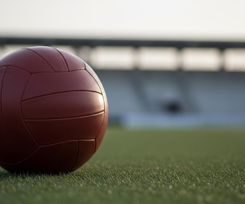 Ciemnoczerwona piłka do siatkówki, wykonana ze sztucznej skóry, leży na zielonej, równej murawie z widocznymi pojedynczymi źdźbłami trawy. Piłka, z klasycznym wzorem paneli i wyraźnymi szwami, jest głównym, ostro sfotografowanym elementem kadru, zajmującym lewą stronę obrazu. W tle, rozmyte i lekko przyciemnione, widać fragment trybun stadionu oraz jasne, niemal białe niebo.