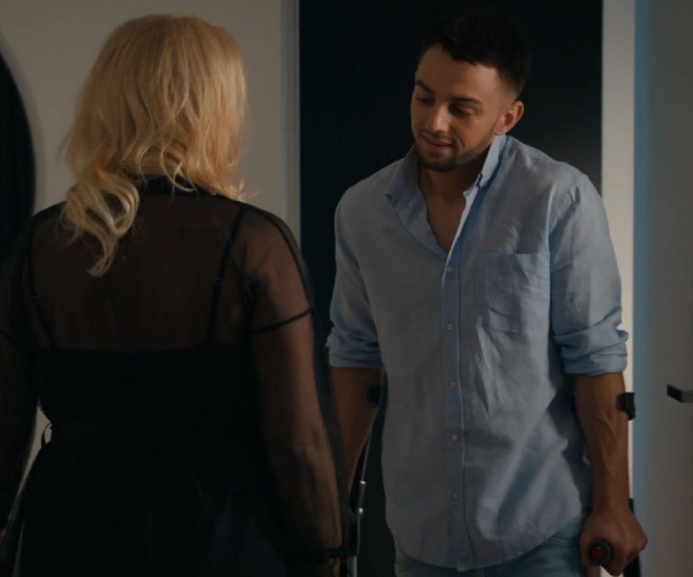 Justyna (Sylwia Juszczak-Krawczyk) odwrócona tyłem, w przezroczystej czarnej narzutce, rozmawia z Mirkiem (Oliwier Kozłowski) w jasnoniebieskiej koszuli. On patrzy na nią, trzymając coś w dłoni. Zdjęcie z serialu Barwy szczęścia o relacji Mirka i Justyny znajdziesz na Super Seriale.