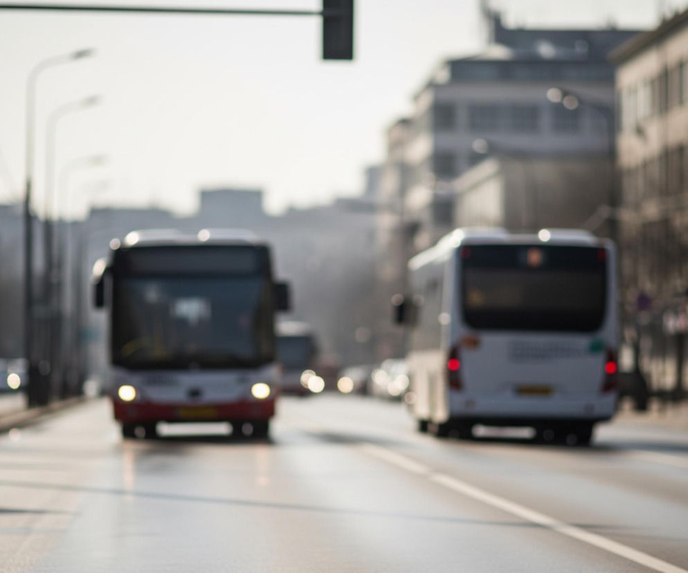 Na zdjęciu widoczna jest rozmyta scena uliczna z dwoma autobusami w centrum, jadącymi w przeciwnych kierunkach. Przedni autobus, z czerwonym pasem u dołu i białymi elementami u góry, zbliża się do widza, mając włączone jasne reflektory. Drugi, biały autobus, oddala się w głąb ulicy, ukazując tył z czerwonymi światłami stop.

Ulica, na której znajdują się pojazdy, jest asfaltowa z białymi liniami rozdzielającymi pasy ruchu. Po obu stronach drogi widać rozmazane budynki, latarnie uliczne i słupy, sugerujące miejski krajobraz. Cała scena jest utrzymana w jasnych, nieco stonowanych barwach, z dużą ilością światła słonecznego rozświetlającego drogę i otoczenie.