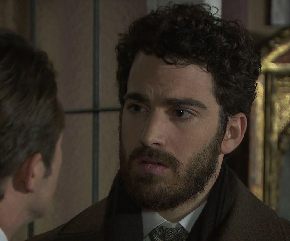 Mężczyzna z ciemnymi, kręconymi włosami i brodą, ubrany w ciemny płaszcz i krawat, patrzy z zamyśloną miną na drugą osobę, której widać tylko tył głowy. Scena pełna dramatyzmu z serialu „Akacjowa 38”, o której więcej przeczytasz na Super Seriale.