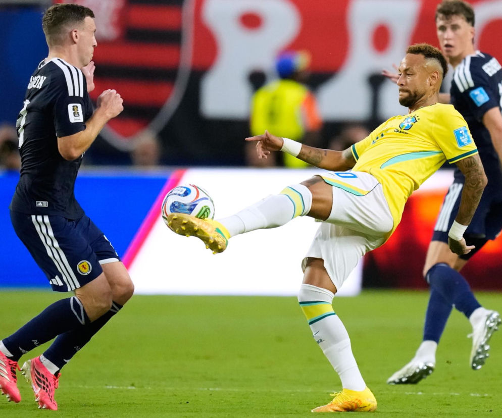Piłkarz Brazylii Neymar w żółtej koszulce uderza piłkę, obok obrońca Szkocji w ciemnym stroju z numerem na plecach. Dynamiczne ujęcie z meczu Brazylia-Szkocja, gdzie Brazylia wygrała 3:0. Więcej o wynikach i przebiegu spotkania przeczytasz na naszym portalu.