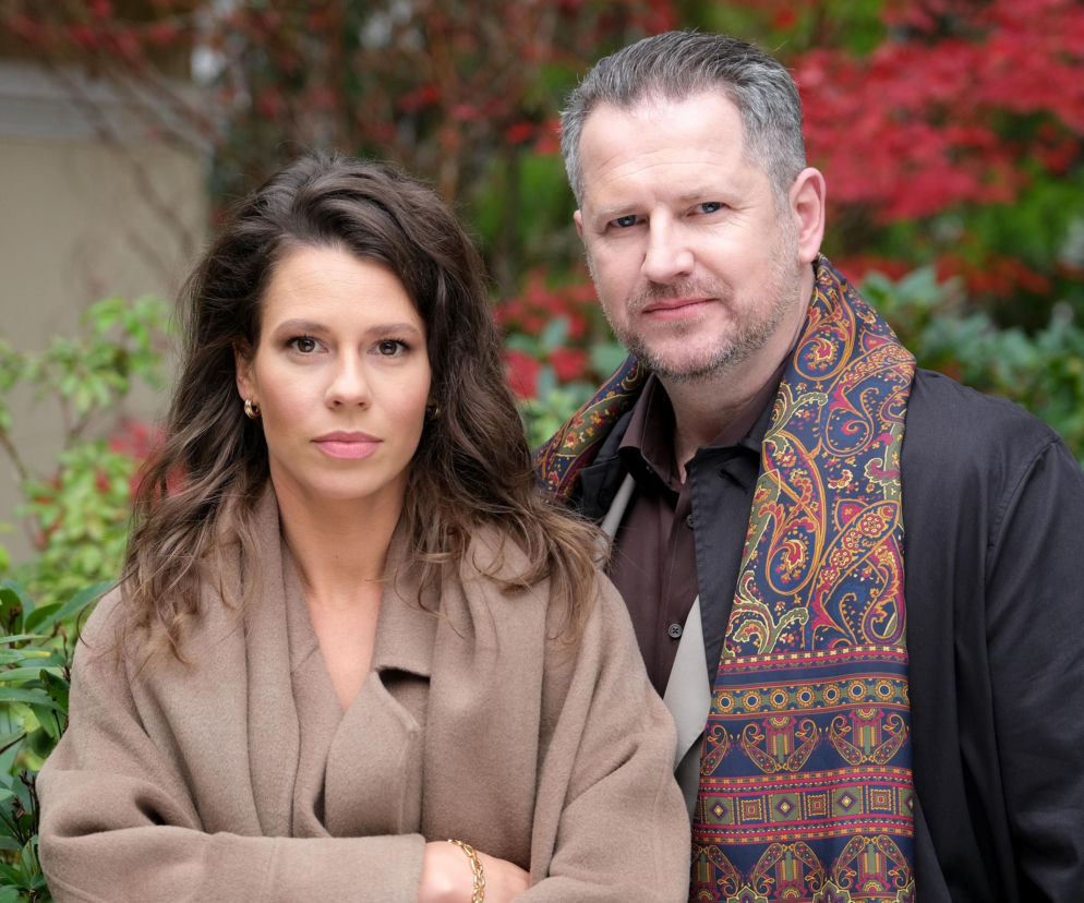 Katarzyna Cichopek i Krystian Wieczorek w stylizacjach z serialu M jak miłość. Aktorka w beżowym płaszczu i ze skrzyżowanymi rękami, aktor w ciemnej koszuli i czarnej kurtce z wzorzystym szalem. W tle rośliny z czerwonymi liśćmi. Więcej o ich losach na Super Seriale.