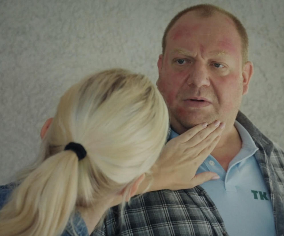 Mężczyzna z czerwonymi plamami na twarzy, Tadeusz (Bartłomiej Nowosielski), oburzony, gdy kobieta z blond włosami w kucyku (Jagoda) delikatnie dotyka jego podbródka. Tadeusz nosi szarą koszulę w kratę nałożoną na jasnoniebieskie polo, widoczny napis TK. Dramatyczna scena z serialu M jak miłość, o której więcej dowiesz się na Super Seriale.