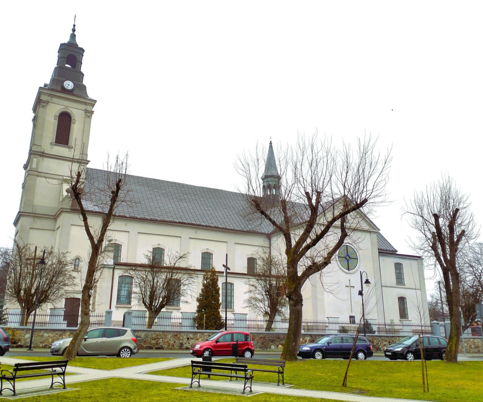 Jasny kościół w Rozprzy z wysoką wieżą i zegarem widziany z perspektywy rynku. O historii miasta czytaj w Eska Łódź.