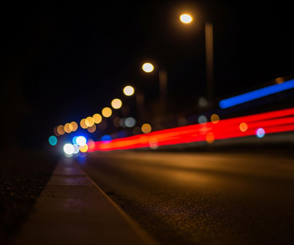 Nocna scena przedstawia miejską ulicę z długimi smugami świateł pojazdów w ruchu. Droga w centralnej części zdjęcia rozciąga się w głąb, a jej asfaltowa nawierzchnia jest ciemna z widocznymi refleksami światła. Po prawej stronie widać dwie wyraźne, poziome smugi: czerwoną na dole i niebieską powyżej, symbolizujące światła tylne i przednie samochodów.

Wzdłuż drogi po lewej stronie biegnie krawężnik, a tuż za nim znajduje się ciemne pobocze. Na horyzoncie, rozciągając się po lewej i górnej części obrazu, widoczny jest rząd rozmytch, okrągłych świateł ulicznych o ciepłej, żółtej barwie, które przypominają efekty bokeh. Tło jest całkowicie ciemne, co potęguje kontrast z jasnymi światłami.