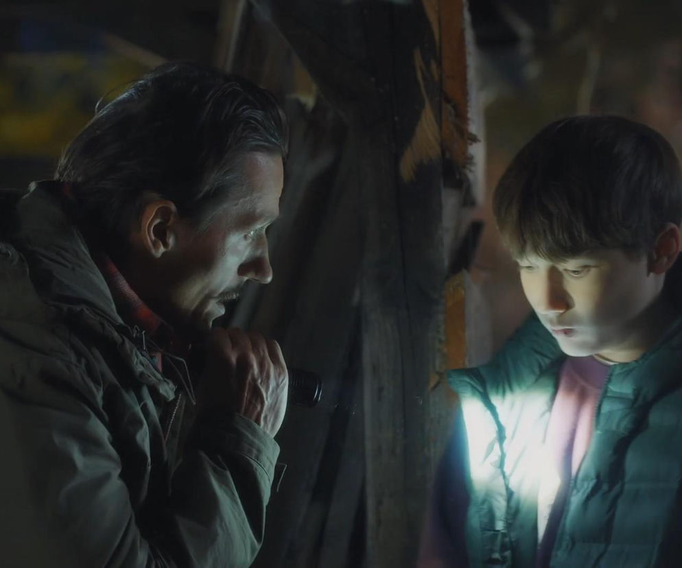 Zastraszony Franek (Jarosław Śmigielski) i bezwzględny ojczym (Marcin Sitek) w scenie z serialu M jak miłość. Mężczyzna trzyma latarkę, a światło pada na twarz chłopca. Więcej o dramacie Franka przeczytasz na portalu Super Seriale.