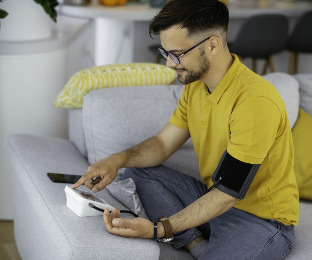 Młody mężczyzna w okularach i żółtej koszulce mierzący ciśnienie krwi w domu, siedzący na szarej kanapie. Obraz symbolizuje rosnący problem nadciśnienia wśród młodych dorosłych, o czym więcej przeczytasz na portalu Poradnik Zdrowie.