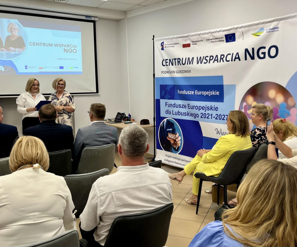 Dwie kobiety na scenie podczas spotkania Centrum Wsparcia NGO Podregion Gorzowski, jedna z nich odbiera dokument. W tle projektor z prezentacją i rollup z informacjami o Funduszach Europejskich dla Lubuskiego. Temat dotacji dla organizacji pozarządowych jest szeroko omawiany na naszym portalu.