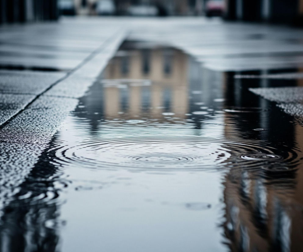 Na mokrej, ciemnej nawierzchni, prawdopodobnie chodniku lub placu, widoczne są kałuże i odbicia. Na pierwszym planie, w centralnej części zdjęcia, znajduje się kałuża z wyraźnymi, koncentrycznymi kręgami na powierzchni wody, sugerującymi upadek kropli. Obok niej, nawierzchnia jest szorstka i pokryta drobnymi, błyszczącymi kropelkami wody, co nadaje jej teksturę.

W odbiciu w kałuży widać zamazane kształty budynków w ciepłych, brązowych i żółtych odcieniach, z pionowymi elementami przypominającymi okna, oraz ciemniejsze pionowe linie w tle. Całe tło jest mocno rozmyte, z niewyraźnymi ciemnymi formami, tworzącymi głębię obrazu. Kolorystyka jest dominująco chłodna, z odcieniami szarości, błękitu i czerni, przełamywana ciepłymi tonami odbicia.