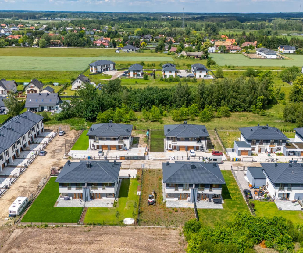 Widok z lotu ptaka na nowe osiedle deweloperskie w Głoskowie, gmina Piaseczno, gdzie szereg nowoczesnych domów jednorodzinnych z ciemnymi dachami sąsiaduje z zielonymi polami i lasami. Ten dynamiczny rozwój budownictwa mieszkaniowego to temat, o którym przeczytasz na Architektura Murator Plus.
