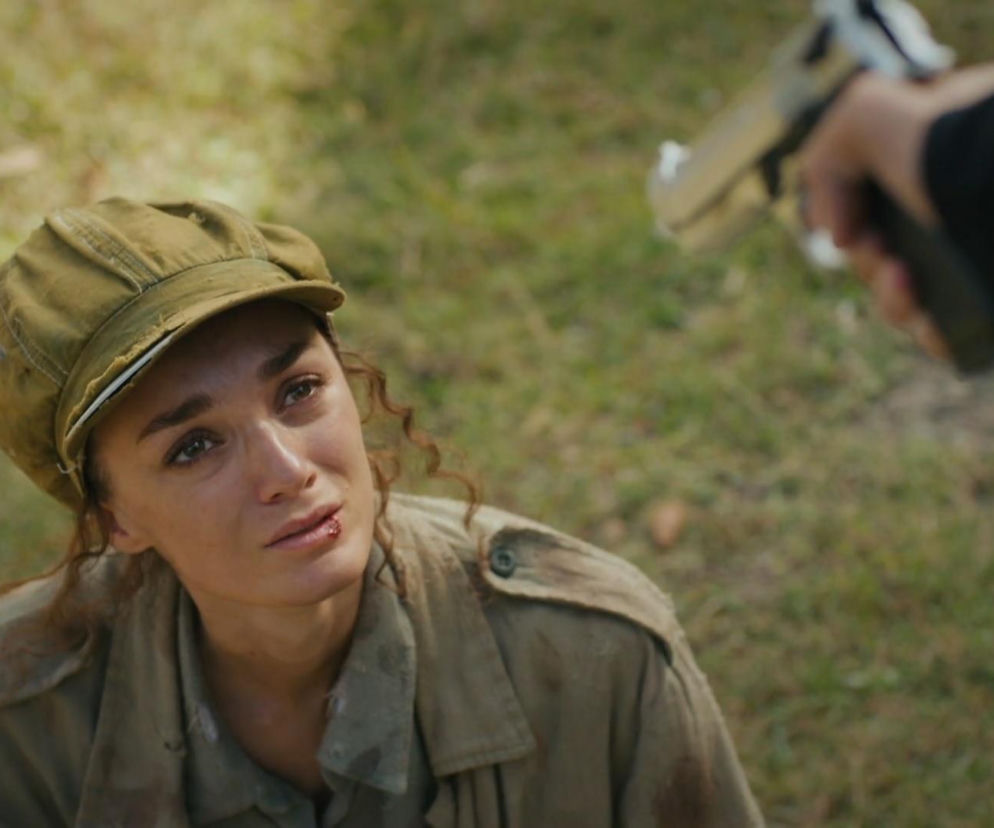 Przestraszona Nana z serialu Dziedzictwo, w brudnym ubraniu i czapce, klęczy na ziemi, a z jej wargi sączy się krew, patrząc z lękiem na lufę pistoletu wycelowaną w jej stronę. Scena budząca emocje, znana z Super Seriale.