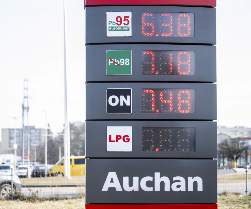 Pylon stacji paliw Auchan z aktualnymi cenami, ukazujący cyfrowe wyświetlacze z kwotami za benzynę Pb95, Pb98, olej napędowy ON oraz LPG. W tle budynki mieszkalne i ulice, co wskazuje na kontekst miejski. O kontrolach cen paliw przeczytasz na Super Biznes.