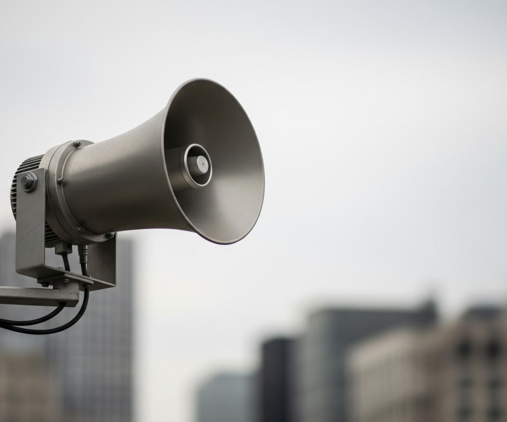 Szary głośnik tubowy, skierowany w prawo, dominuje w lewej części kadru. Jest on zamocowany na metalowym wysięgniku, od którego odchodzą czarne kable. W tle widać rozmyte, szare sylwetki budynków, a za nimi jasne, zachmurzone niebo.