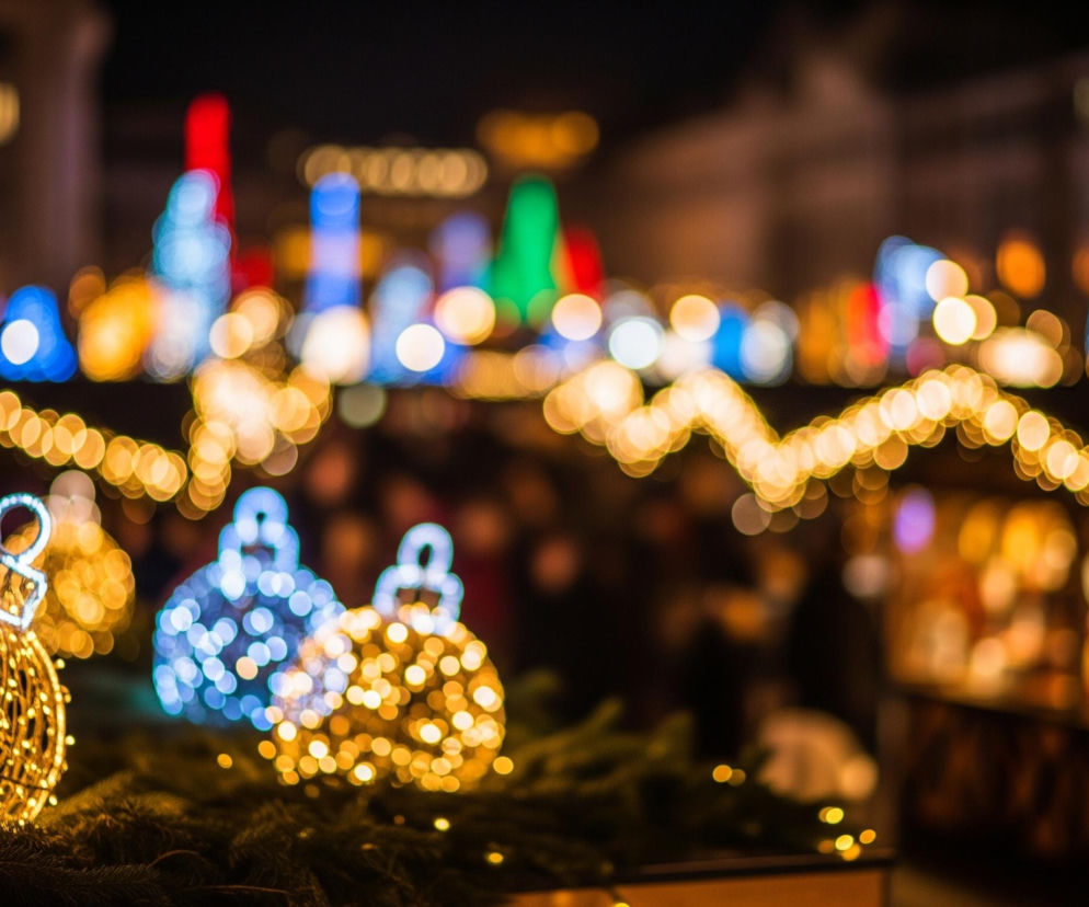 Na pierwszym planie, z lewej strony, widać fragmenty zielonych gałęzi iglastych drzew, udekorowanych małymi, świecącymi na żółto lampkami, tworzącymi kształt serca. Centralnie umieszczona jest duża, złota bombka świąteczna, wykonana z siatki drucianej i oświetlona od wewnątrz drobnymi, ciepłymi lampkami. Za nią, po prawej stronie, znajdują się dwie podobne, rozmyte bombki: jedna w odcieniach błękitu i bieli, druga w żółto-pomarańczowych barwach. Tło stanowi rozmyty krajobraz miejski w nocy, z licznymi, kolorowymi światłami w odcieniach czerwieni, zieleni, błękitu i żółci, sugerującymi jarmark lub świąteczne dekoracje.