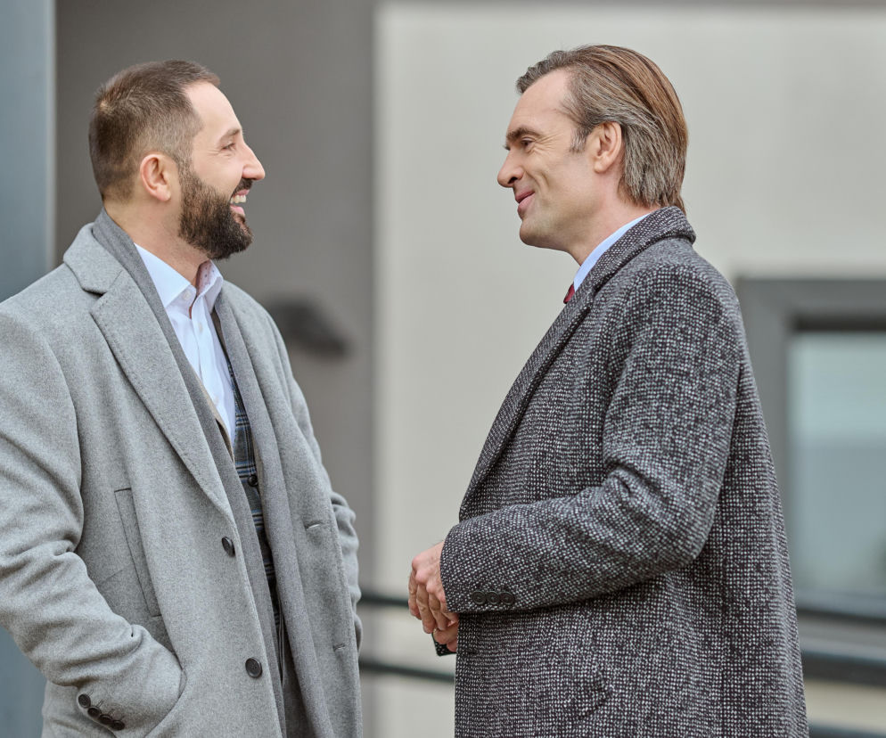 Dwóch elegancko ubranych mężczyzn, w szarych płaszczach, uśmiecha się do siebie w trakcie rozmowy na tle nowoczesnego budynku. Ich postacie w scenie z serialu Na Wspólnej znajdziesz na Super Seriale.