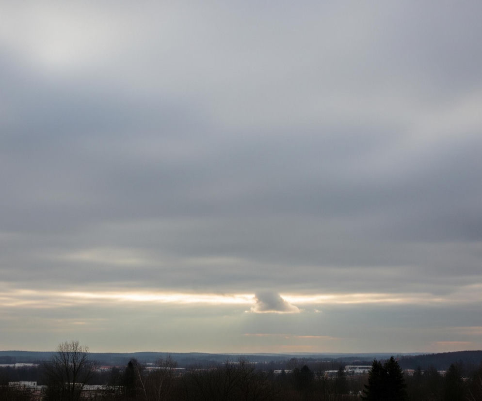 Szeroki plan nieba pokrytego szarymi chmurami, z jasnym, poziomym pasmem światła prześwitującym przez chmury w centralnej części. Na tym pasie światła, nieco na prawo od środka, znajduje się mała, puszysta chmura, która blokuje fragment blasku. Poniżej horyzontu rozciąga się ciemny krajobraz z widocznymi zarysami wzgórz i drzew pozbawionych liści, oraz kilkoma białymi plamami, które mogą być śniegiem lub budynkami.