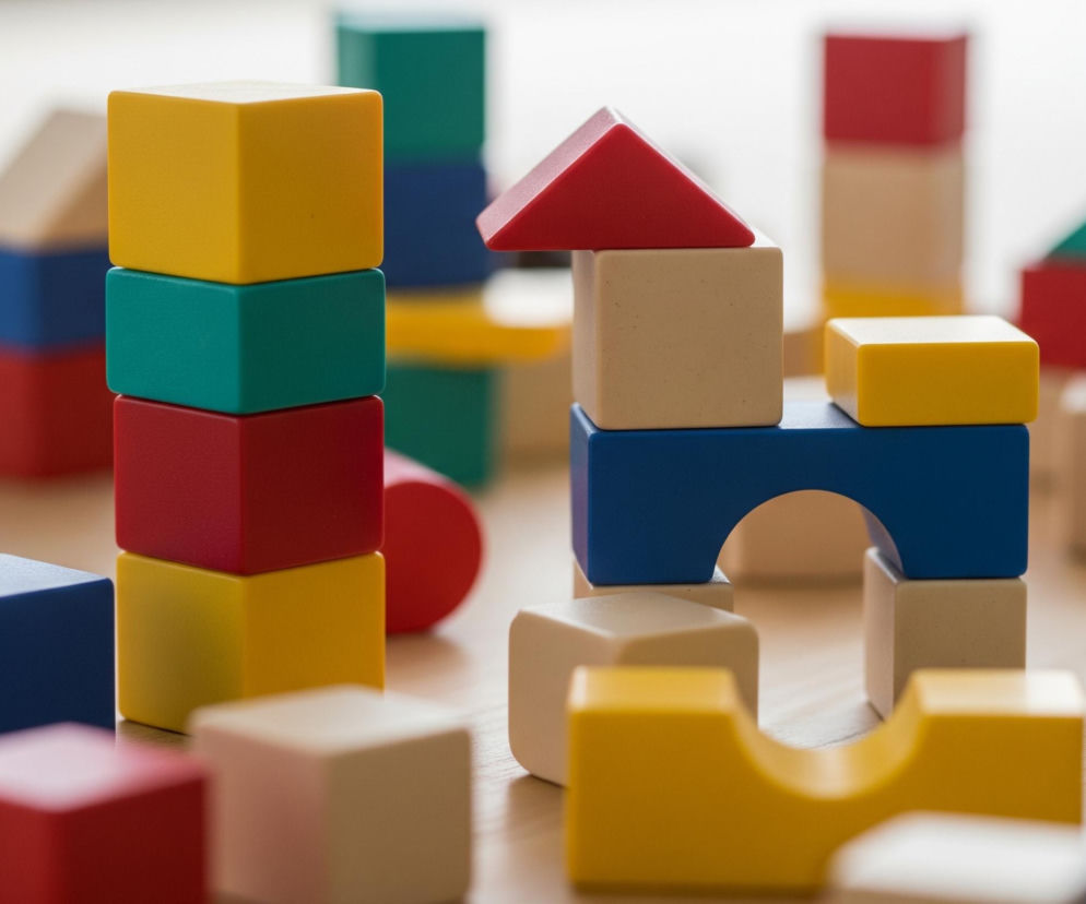 Rozrzucone klocki konstrukcyjne na jasnym, drewnianym podłożu. Centralnie położona jest wieża zbudowana z czterech klocków: żółtego, turkusowego, czerwonego i ponownie żółtego na dole. Obok niej znajduje się inna konstrukcja składająca się z beżowego sześcianu i niebieskiego klocka z otworem w kształcie łuku, na którym leży żółty prostopadłościan, a wszystko to wieńczy czerwony dach w kształcie trójkąta. Cała scena jest rozmyta z tyłu, z wyraźnym pierwszym planem, ukazującym różnorodność kształtów i kolorów klocków, w tym niebieskie sześciany, beżowe prostopadłościany, czerwone i żółte elementy, a także turkusowy klocek z wypustkami.